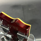 Close-up of the headstock of a guitar, showing a glossy dark red/burgundy finish with a yellow binding edge and silver tuning pegs against a dark gray background.