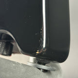 Close-up of a glossy black guitar body corner with visible wear, scuffs, and a small gold-toned mark near the edge, mounted above a silver hardware piece.