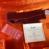 A Fender guitar strap in dark reddish-brown leather, displayed with its Certificate of Authenticity and a small clear bag containing pink accessories on a textured orange-brown fabric background.
