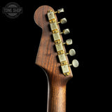 Close-up of a guitar headstock with wooden finish and gold tuning pegs on a black background, featuring 'Tone Shop' logo.