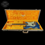 A blue electric guitar resting inside an open silver hard case with bright orange textured lining, shown against a black background. A Tone Shop watermark is visible in the top-left corner.