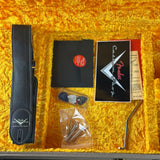Guitar case contents arranged in a yellow plush-lined case, including a black leather strap, black envelope-style pouch with a red circular sticker, printed cards, a small accessory in a clear bag, and a metal tool.