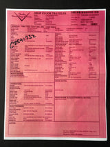 A pink paper form labeled 'SHOP FLOORS TRAVELLER' with dense printed text, a barcode, and a handwritten black ink signature, photographed against a white background.