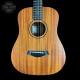 A close-up of a wooden acoustic guitar body with a warm honey-toned wood grain finish, black rosette, and black bridge, shown against a solid black background. A 'TONE SHOP' watermark is visible in the top-left corner.