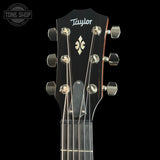 Close-up of a Taylor acoustic guitar headstock against a black background, showing six silver tuning pegs, six strings, and the Taylor logo.