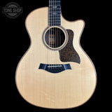A Taylor acoustic-electric guitar with a natural wood finish and dark pickguard, shown against a solid black background with a Tone Shop watermark in the top left corner.