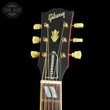 Close-up of a Gibson humbucker guitar headstock with six gold tuning pegs, a black face, and the Gibson logo in gold script, shown against a black background.