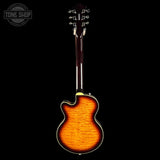 A back view of an electric guitar with a vibrant sunburst finish showing orange and brown wood grain, set against a solid black background with a Tone Shop logo in the top left corner.