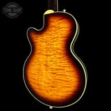 Close-up of the back of a double-cutaway electric guitar showing a vibrant flamed maple top with a classic sunburst finish, transitioning from bright amber-orange in the center to dark brown and black at the edges, set against a solid black background.