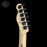 Close-up of the headstock of a natural wood electric guitar against a solid black background, featuring six black tuning pegs and a Tone Shop watermark in the upper left corner.