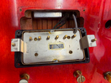 Close-up of a guitar humbucker pickup component mounted on a bright red guitar body, showing the metal pole pieces, screws, and wiring.