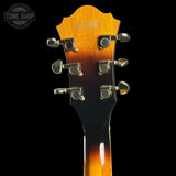 Close-up of the headstock of an acoustic guitar against a black background, showing six silver tuning pegs and a sunburst finish transitioning from amber-orange to black. A Tone Shop watermark is visible in the top-left corner.
