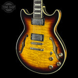 A classic electric guitar with a natural flame maple top showing a sunburst finish that transitions from bright yellow-orange in the center to dark brown and black at the edges. It features gold-toned hardware, black pickups, and white binding on a solid black background.