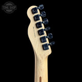 Close-up of the headstock of a Fender electric guitar in natural wood finish with black tuning pegs, shown against a solid black background. A Tone Shop watermark is visible in the top left corner.
