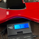 A close-up of a red electric guitar body resting on a digital smart weigh scale displaying 7.12 kg, set on a dark wooden surface.