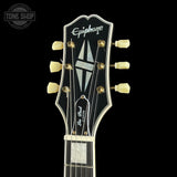 Close-up of the headstock of an Epiphone Les Paul Custom electric guitar against a solid black background, showing the gold tuners and white brand lettering.
