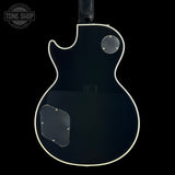 The back view of a black electric guitar body with white binding and clear protective decals, shown against a solid black background with a Tone Shop watermark in the top left corner.