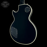 The back of a black electric guitar with a white binding edge, shown against a solid black background. A circular 'TONE SHOP' watermark is visible in the top-left corner.