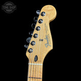 Close-up of a Fender Stratocaster electric guitar headstock in natural maple wood, showing six black tuning pegs and the Fender logo, set against a solid black background.