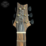 Close-up of a custom electric guitar headstock and neck, featuring a dark wood grain finish, six black tuning pegs, and a white script signature on the headstock, set against a solid black background.