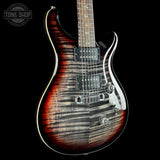 A close-up studio shot of an electric guitar body against a black background, showing a sunburst flame maple finish that transitions from dark brown to black and reddish amber. The guitar features chrome hardware, a black pickguard, and a white binding edge.