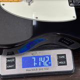 A close-up of a digital scale displaying 7.142 lbs on a dark wood surface, positioned under the black body and cream pickguard of an electric guitar.