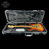An electric guitar with a flame maple body in a sunburst finish, displayed inside an open silver hard case with a black velvet interior. The brand headstock is visible on the neck.