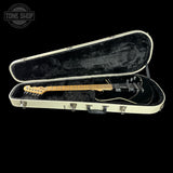 An electric guitar with a black body and natural wood neck resting inside an open cream-colored hard case with black plush lining, shown against a solid black background.