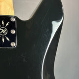 Close-up of a black electric guitar body with a glossy finish, showing a black pickguard with a white stylized 'R' logo and silver screws, along with part of the natural wood neck.