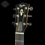 Close-up of the headstock of a Taylor acoustic guitar against a black background, showing the brand name Taylor in white script, a gold treble treble logo, and six silver tuning pegs.