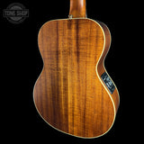 Close-up of the back of a wooden acoustic guitar against a black background, showing the rich grain of the wood and a Tone Shop logo watermark in the top left corner.