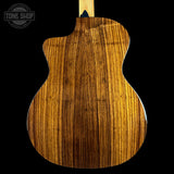 Close-up of the back of an acoustic guitar showing a rich, glossy wood grain finish in warm brown and golden tones, set against a solid black background with a Tone Shop watermark in the top left corner.
