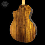 Close-up of the back of an acoustic-electric guitar showing a rich, warm brown wood grain finish on the body, with a black background and a Tone Shop watermark in the top left corner.