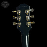 Close-up of the headstock of a black electric guitar with gold-toned tuning pegs, shown against a solid black background. A Tone Shop watermark is visible in the top left corner.