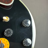 Close-up of a black electric guitar body showing part of the bridge area, control knobs, a yellow volume knob, and a white edge trim against a dark gray background.