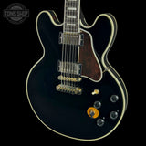 A black electric guitar with cream binding, a dark wood neck, gold hardware, and black control knobs, shown against a black background with a Tone Shop watermark in the top left corner.