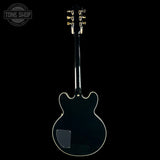 A black electric guitar shown from the back against a solid black background, featuring gold tuning keys and a white binding edge.