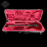 A red electric guitar resting inside an open hard case with bright pink velvet lining, photographed against a black background. A Tone Shop watermark is visible in the top-left corner.