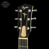 Close-up of a Taylor acoustic guitar headstock with six silver tuning pegs, a black glossy finish, gold script branding, and a natural wood bridge plate, shown against a black background.