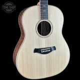 An acoustic guitar with a natural light wood finish and dark brown sides, shown against a solid black background. A circular 'TONE SHOP' watermark is visible in the top-left corner.