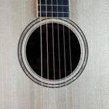 Close-up of an acoustic guitar body showing the circular sound hole, rosette, six strings, and part of the light wood neck against a pale wood-grain background.