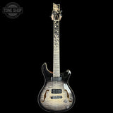 A double-cutaway electric guitar with a natural wood grain finish, shown vertically against a solid black background. The headstock features a decorative gold-toned logo, and the neck has a white maple fretboard with black inlays.