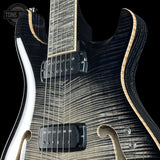 Close-up of an electric guitar body showing a flamed maple top with a black and silver woodgrain finish, black humbucker pickups, and a light natural wood edge, set against a black background.