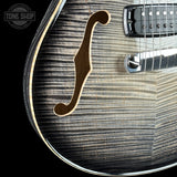 Close-up of an electric guitar body showing a black arched top with a natural wood f-hole, a black pickup, and a silver tremolo arm against a black background. A Tone Shop watermark is visible in the top left corner.