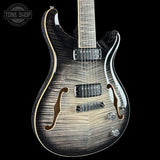 A close-up of an electric guitar body against a black background, featuring a natural flame maple wood grain finish that transitions from dark brown to light beige. The guitar has gold-toned binding, dual black humbucker pickups, and a chrome bridge.