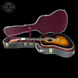 An acoustic guitar with a sunburst finish displayed inside an open hard case with a burgundy velvet interior, set against a solid black background.