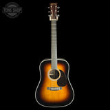 A vintage-style acoustic guitar with a sunburst finish, shown vertically against a solid black background. The guitar features a dark brown headstock with gold-toned tuning pegs, a black pickguard, and a white binding around the body.