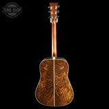 The back of an acoustic guitar with a rich, swirling natural wood grain pattern on the backplate, a matching wood neck, and silver tuning keys, shown against a solid black background with a Tone Shop watermark in the top left corner.