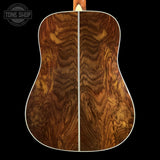 Close-up of the back of an acoustic guitar showing a rich, symmetrical wavy wood grain pattern in warm brown and golden tones, with a white binding edge and a TONE SHOP logo in the top left corner.