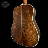Close-up of the back of an acoustic guitar showing a rich, highly figured brown wood top with a white center stripe and white edge binding, set against a solid black background.